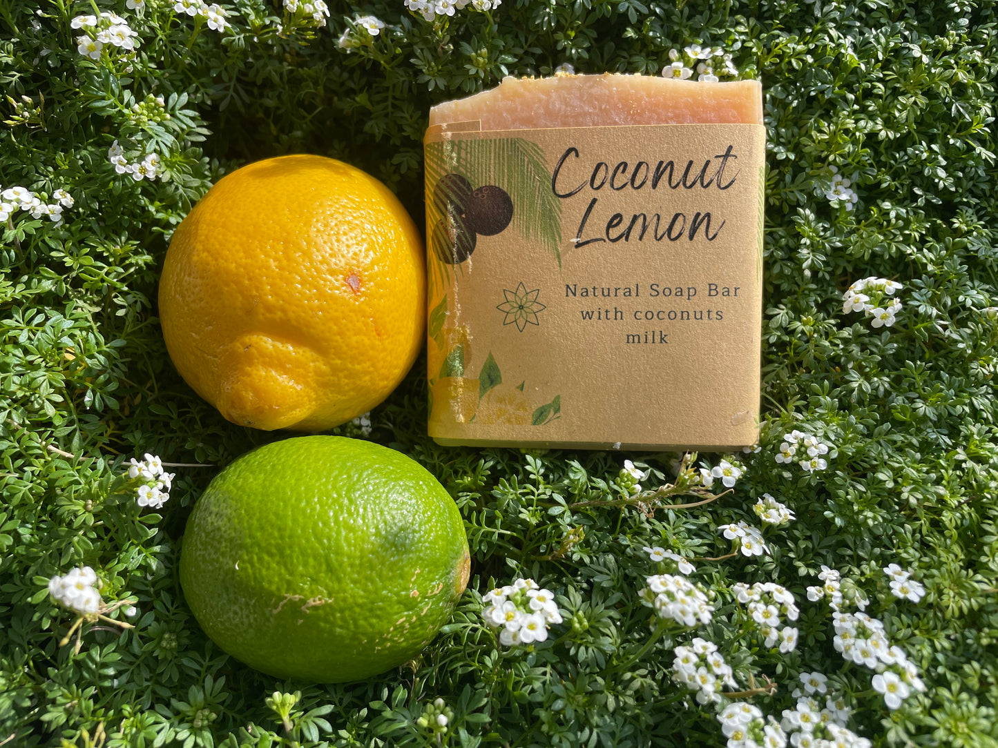 Coconut Lemon soap with real coconut milk, scented with coconut and lemon essential oil, the vegan soap is showing the front label and on the side it has 2 lemons green and yellow, everything is lay on a bed of wild flowers