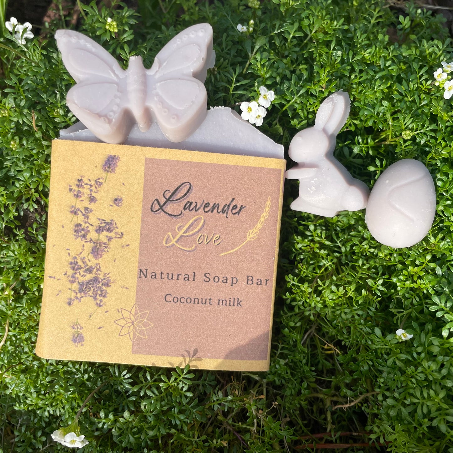 Lavender love natural handcrafted vegan soap, natural coloured, with a purple pastel tone, made with coconut milk, and relaxing lavender essential oil. the photo show the front side of the soap bar and 3 samples in the shape of a butterfly, a bunny and an easter egg, all on a bed of wild flowers